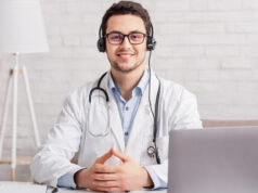 Health blog. Young doctor in headphones sitting at table with laptop